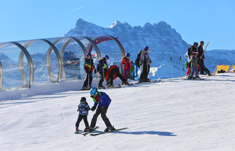 Kind lernt Skifahren im White Valley mit einem Erwachsenen. Im Hintergrund ein überdachtes Förderband mit einer Gruppe von Menschen am Ende. Dents du Midi im Hintergrund.
