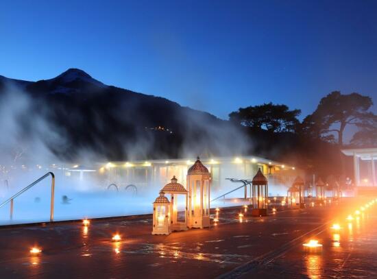 La nuit tombe, la montagne et des arbres se découpent en ombre chinoise dans le ciel, derrière le bassin embrumé.
Au premier plan, plusieurs lanternes et bougies allumées.