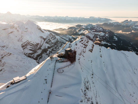 A bird’s-eye view of the Glacier 3000 summit station at Scex Rouge, looking out over the snow-covered glacier and the valley in the background. The photo was taken at the end of the day in warm, low-angle light, with the new station shown in black